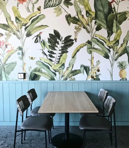 Mural botaniczny Tropicana na ścianie z lamperią i stołem z krzesłami.webp
